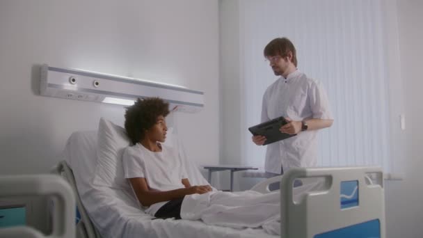 手のタブレットを持つ医師は アフリカ系アメリカ人のティーンエイジャーと手術結果を議論します 看護師はベッドに横たわる少年たちの足を包帯している 成功した手術後に回復する患者 明るい病院の部屋 — ストック動画