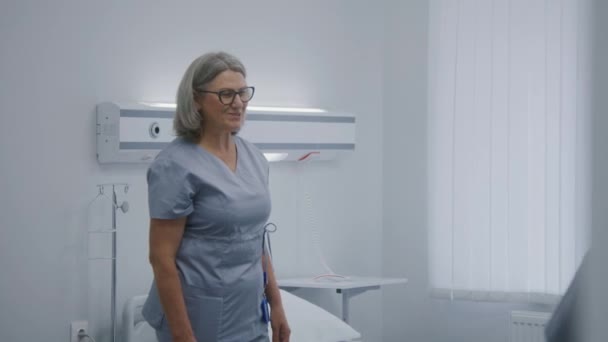 成熟的护士在医院房间里用遥控装置给病人铺床 女性医务工作者推动轮椅与老年妇女 把她放在床上 现代诊所或医疗设施 慢动作 — 图库视频影像