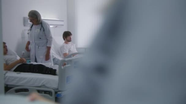 护士拿出午餐盒 交给卧床休息的病人 成熟的女医生对病人进行检查 成年男子在手术成功后完全康复 医护人员在医院病房工作 — 图库视频影像