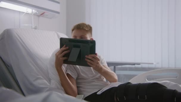 Mladý Chlapec Obvázanou Nohou Leží Posteli Mluví Tátou Pomocí Tabletu — Stock video