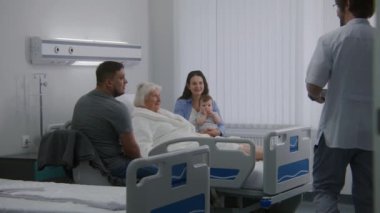 Yaşlı kadın hasta hastane koğuşunda yatıyor ve ailesiyle vakit geçiriyor. Doktor dijital tablet kullanır, başarılı bir ameliyattan sonra iyileşen yaşlı kadınla konuşur. Modern tıbbi tesis. Yavaş çekim.