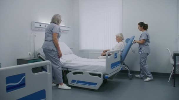 成熟した看護師は リモートコントロールを使用してベッドをセットアップし 患者のための病院室を準備します 女性の医療従事者は 高齢女性と車椅子を押し ベッドに入れます 現代のクリニックや医療施設 — ストック動画