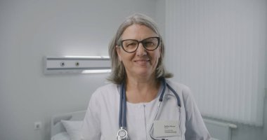 Yetişkin bir kadın doktor kameraya bakar ve gülümser. Boynunda steteskop olan profesyonel bir sağlık uzmanı hastane koğuşunun ortasında duruyor. Modern tıp merkezinde çalışan tıbbi personel.