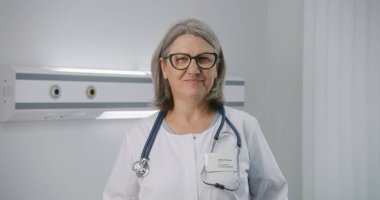 Boynunda steteskop olan profesyonel kadın doktor hastane odasının ortasında duruyor. Olgun doktor kameraya bakar, gülümser ve gözlükleri ayarlar. Modern tıp merkezinde tıbbi personel çalışıyor..