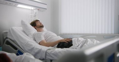 Bayan hemşire hasta yatağında dinlenmeye gelir, kalp atış hızı monitörünü parmağına takar ve serumu düzeltir. Adam başarılı bir ameliyattan sonra tamamen iyileşiyor. Hastane koğuşunda tıbbi personel.