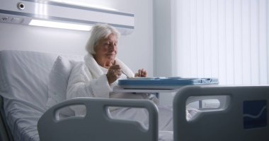 Yaşlı kadın lezzetli yemekler yiyor ve parlak hastane koğuşundaki yatakta dinlenen kameraya bakıyor. Başarılı bir ameliyattan sonra iyileşen yaşlı kadın hasta. Modern tıbbi tesis ya da klinik. Portre.
