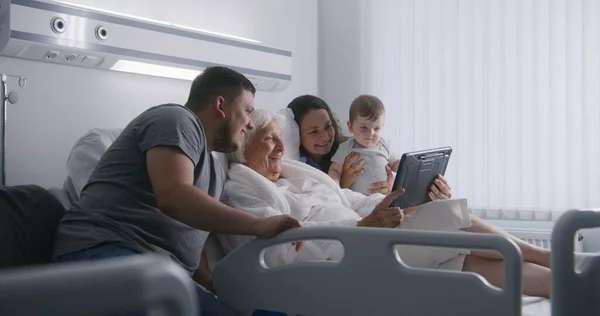 Hospital room. Loving family support elderly grandmother recovering after successful surgery. Relatives talk by video call using digital tablet, spend time together. Modern medical facility, clinic.