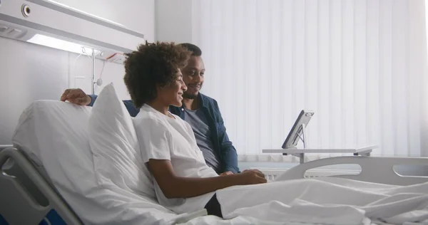 アフリカ系アメリカ人のティーンエイジャーは 病院の部屋でベッドに父親と一緒に横たわっています 家族はタブレットのコンピュータを使用して遠隔で話します 患者は 手術が成功した後に完全に回復します クリニックで働く医療関係者 — ストック写真