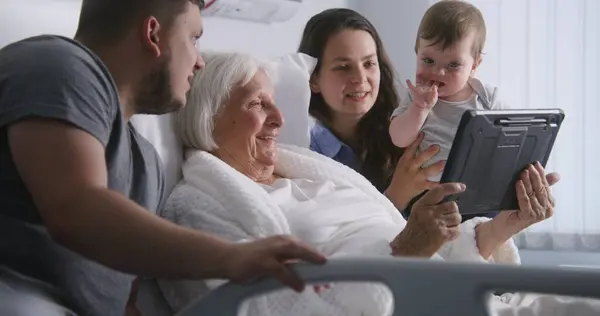 病院の部屋 ベッドに横たわる老婦人は親戚と時間を過ごす 家族を愛するタブレットコンピュータを使用し 手術が成功した後に回復する高齢のおばあちゃんをサポートします 現代の医療施設やクリニック — ストック写真