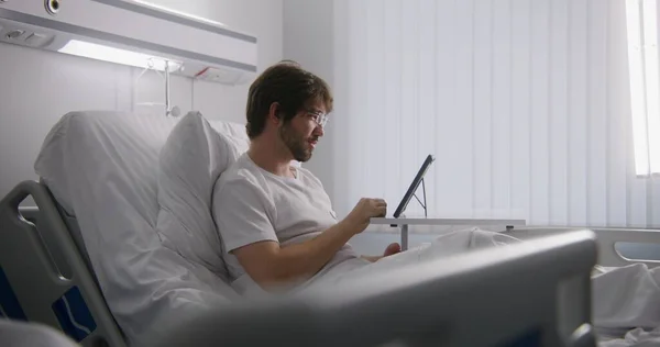 大人の男性は病院の病棟のベッドに横たわり タブレットを使用してオンライン会議でパートナーと話します メガネを着ている男性は 遠隔操作で働きます 成功した手術後に完全に回復する患者 現代明るいクリニック — ストック写真