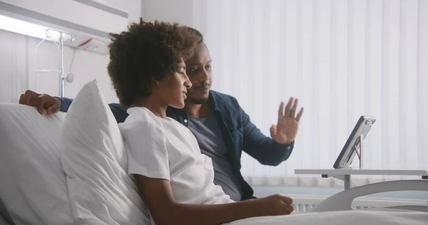 アフリカ系アメリカ人のティーンエイジャーは 病院の病棟でベッドで休む 男は手術後に息子を訪ねている 家族はタブレットのコンピュータを使用して遠隔で話します 患者は 手術が成功した後に完全に回復します 現代クリニック — ストック写真