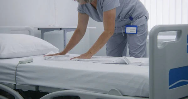 Mature cleaner changes bedclothes and puts clean pillow on bed. Nurse makes beds in hospital ward and prepares it for new patients in bright hospital ward. Medical staff work in modern medical center.