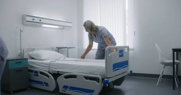 成熟したクリーナーは布を変更し ベッドにきれいな枕を置きます 看護師は病院の病棟でベッドを作り 明るい病院病棟の新しい患者に備えています 現代医療センターで働く医療スタッフ — ストック写真