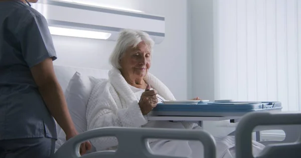 看護師は 女性の老人の食事と大きな弁当箱を開けます 高齢の女性は明るい病院の部屋でベッドで眠り 手術が成功した後に回復し 食べ物を食べました 現代の医療施設やクリニック — ストック写真