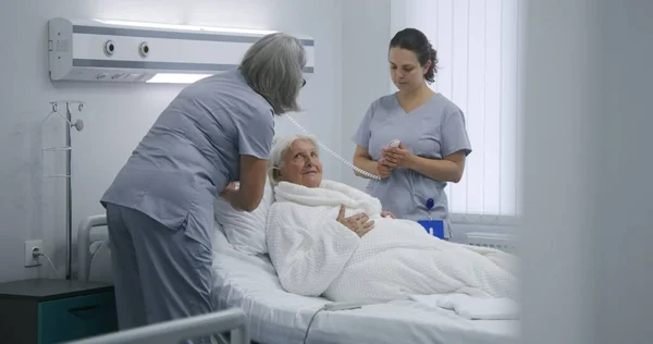 成熟した看護師は リモートコントロールを使用してベッドをセットアップし 患者のための病院室を準備します 女性の医療従事者は 高齢女性と車椅子を押し ベッドに入れます 現代のクリニックや医療施設 — ストック写真
