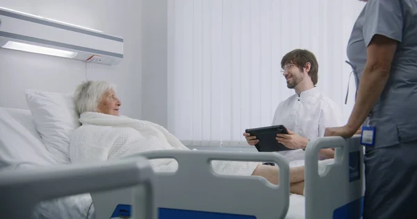 Dokter Consulteert Vrouwelijke Oudere Patiënt Liggend Rustend Bed Ziekenhuiskamer Mannelijke — Stockfoto