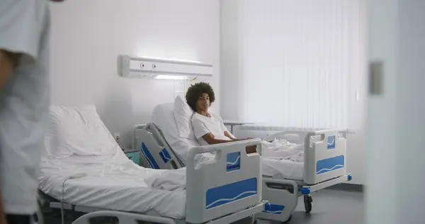 包帯された足を持つ少年は病院の病棟に入ってきて アフリカ系アメリカ人のルームメイトと挨拶する 医療スタッフが患者を診察する 成功した作戦後に回復する人々 現代明るいクリニック — ストック写真