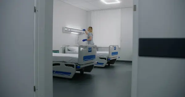 Adult nurse mops floor between beds in hospital ward. Health worker brings cleaning trolley to hospital room. Female cleaner prepares ward for new patients. Medical staff at work in modern clinic.