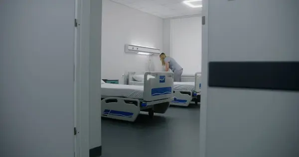 Adult nurse mops floor between beds in hospital ward. Health worker brings cleaning trolley to hospital room. Female cleaner prepares ward for new patients. Medical staff at work in modern clinic.