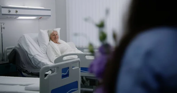 高齢の女性患者は 明るい病院区のベッドに横たわり 眠っている 愛する家族のメンバーは 手術が成功した後に回復する高齢女性に来て 花を与えます 現代の医療施設やクリニック — ストック写真