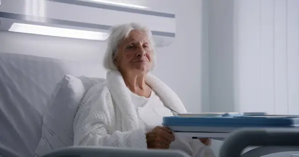 高齢の女性は美味しい食べ物を食べて 明るい病院区のベッドで眠っているカメラを見ています 成功した手術後に回復する女性の高齢患者 現代の医療施設やクリニック ポートレイト — ストック写真