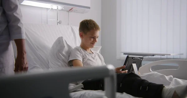 Young boy with bandaged leg lies on bed, talks with dad using tablet. Kid remotely shows hospital conditions to father. Patient fully recovering after successful surgery. Modern and bright clinic.