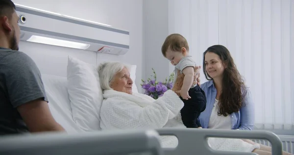 高齢の女性患者は 明るい病院区のベッドに横たわり 小さな孫と遊んでいます 愛する家族のメンバーは 手術が成功した後に回復する上級女性をサポートしています 現代医療施設について — ストック写真