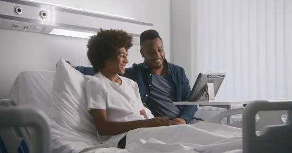 アフリカ系アメリカ人のティーンエイジャーは 病院の部屋でベッドに父親と一緒に横たわっています 家族はタブレットのコンピュータを使用して遠隔で話します 患者は 手術が成功した後に完全に回復します クリニックで働く医療関係者 — ストック写真
