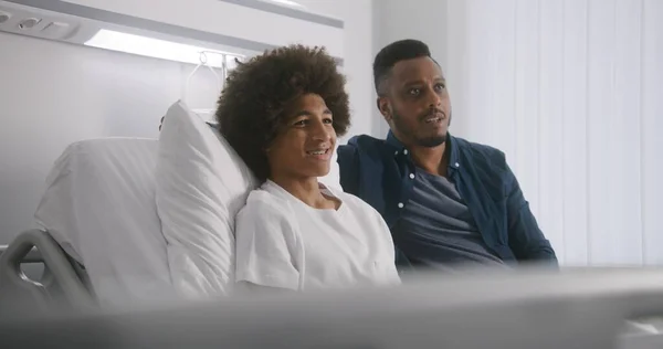 Happy African American teenager rests in hospital room and watches movie together with father. Roommates discuss film. Patient fully recovering after successful surgery. Modern bright medical center.