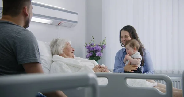 高齢の女性患者は 明るい病院区のベッドに横たわり 孫と娘と話しています 愛する家族は 手術が成功した後に回復する上級女性をサポートします 現代の医療施設やクリニック — ストック写真
