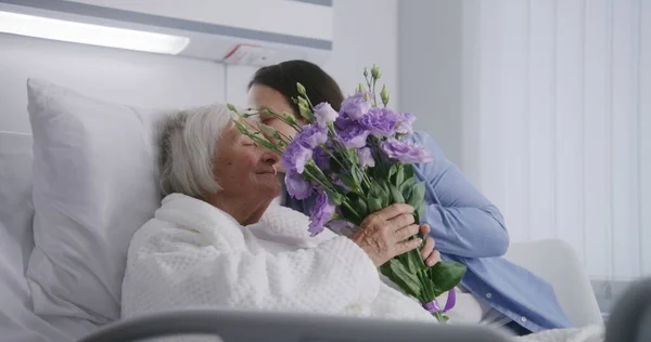小さな子供を持つ愛する家族は 手術が成功した後に回復する祖母を訪れ 花を与えます 高齢の女性は嘘をつき 明るい病院の病棟で寝ている 現代の医療施設やクリニック — ストック写真