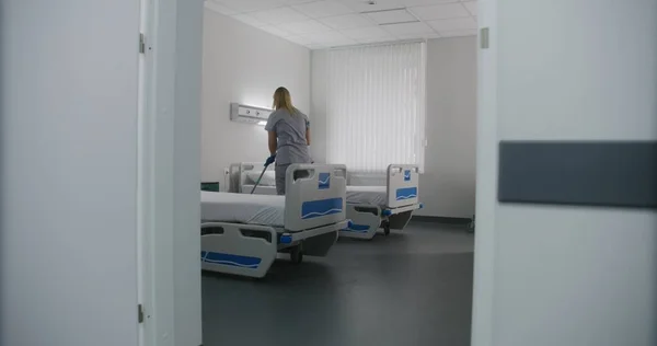 大人の看護師は 病院区のベッドの間の床をモップします 医療従事者はトロリーを病院の部屋に掃除する 女性のクリーナーは新しい患者のための病棟を準備します 近代クリニックで働く医療スタッフ — ストック写真