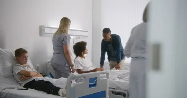 成熟した医師は若い男の子の足を包帯します アフリカ系アメリカ人が病院の病棟で息子を訪ねる 看護師がティーンエイジャーに来て ベッドから立ち上がる 正常な操作の後で完全に回復する患者 — ストック写真