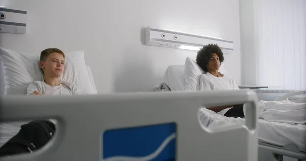 アフリカ系アメリカ人のティーンエイジャーがルームメイトと映画について話している 包帯された足を持つ少年は 病院の部屋でベッドに寝ている 患者は 手術が成功した後に完全に回復します 現代明るい医療センター — ストック写真