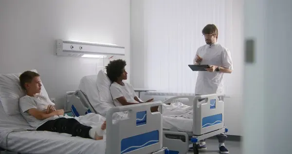 手握平板电脑的医生与非洲裔美国青少年讨论手术结果 躺在床上的小男孩的护士绷带腿 手术成功后病人康复 明亮的病房 — 图库照片