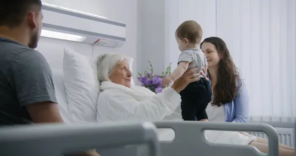 高齢の女性患者は 明るい病院区のベッドに横たわり 小さな孫と遊んでいます 愛する家族のメンバーは 手術が成功した後に回復する上級女性をサポートしています 現代医療施設について — ストック写真