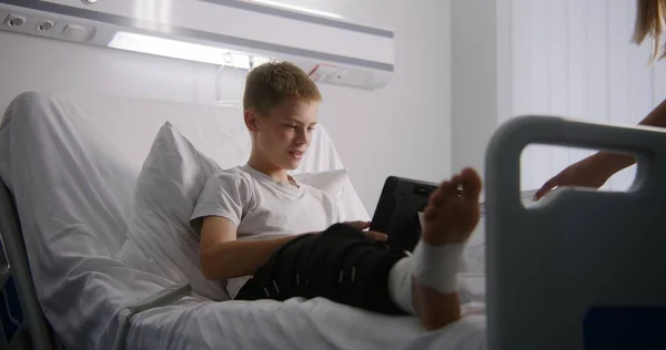 穿着绷带腿的小男孩躺在床上 和朋友们在网络游戏中聊天 医院病房的孩子们在玩平板电脑游戏 手术成功后 病人完全康复 现代明亮的诊所 — 图库照片