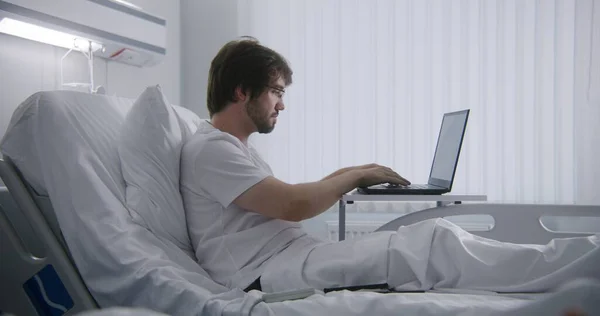 Adult man lies on bed in hospital ward, remotely works using laptop. Programmer works in medical center and chats using phone. Patient fully recovering after successful surgery. Modern bright clinic.