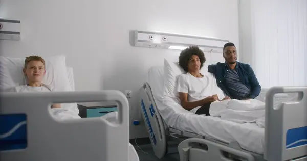 看護婦はベッドの子供に横たわる足を包帯している アフリカ系アメリカ人の少年は病院の病棟で休憩し 父親と一緒にテレビを見ている 成功した手術後に回復する患者 現代明るい医療センター — ストック写真