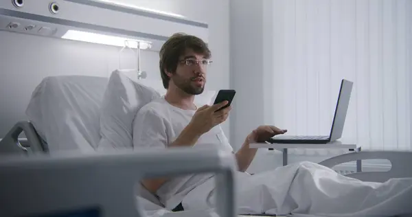 成年男子躺在医院病房的床上 用笔记本电脑远程工作 程序员在医疗中心工作 用电话聊天 手术成功后病人完全康复 现代明亮的诊所 — 图库照片