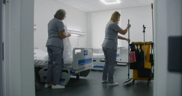 Health worker brings cleaning trolley to hospital ward. Nurses clean hospital room after patients. Mature cleaner with her colleague changes bedding on beds. Medical staff at work in modern clinic.