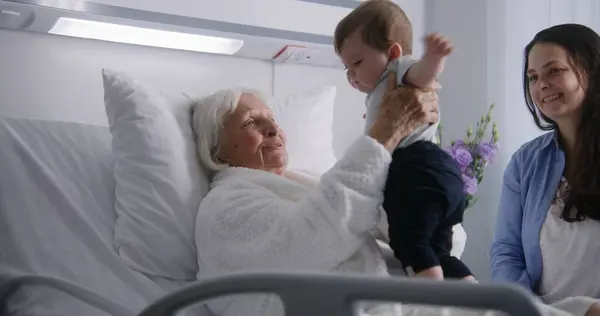 老年女性病人躺在明亮的病房的床上 和小孙子玩耍 有爱心的家庭成员支持老年妇女在手术成功后康复 现代医疗设施 — 图库照片