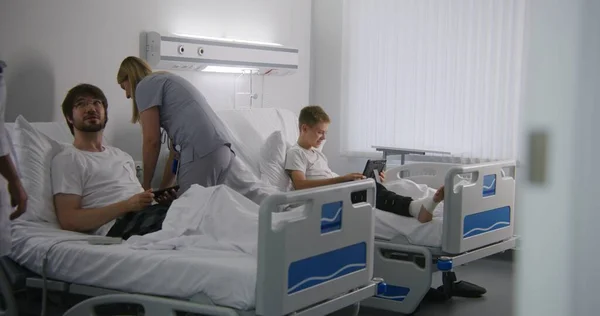 包帯された足を持つ少年はベッドの上に横たわり タブレットを使用して両親と話します 女性医師は ベッドコントローラーの使い方を説明します 成功した手術後に完全に回復する患者 現代病院 — ストック写真