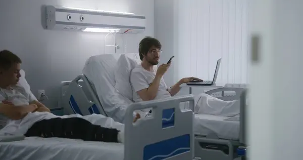 大人の男性は病院の病棟でベッドに横たわり 電話を使って顧客とチャットします メガネの男は遠隔医療センターで働いています 患者は 手術が成功した後に完全に回復します 現代明るいクリニック — ストック写真