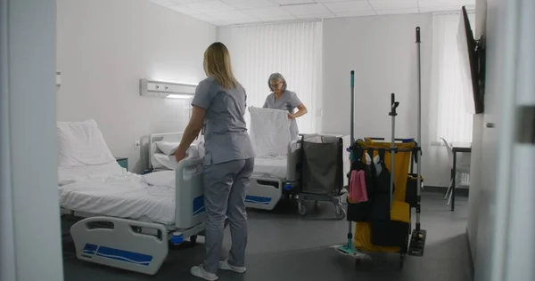 Adult cleaner mops floor with broom in hospital room. Mature health worker changes bedclothes after patients. Nurses clean hospital ward and talk. Medical staff at work in modern medical center.