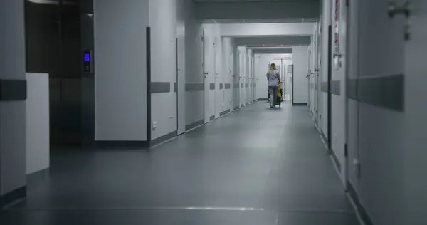 Adult nurse pushes cleaning trolley down medical center hallway. Health worker goes to hospital ward for cleaning. Cleaner takes care of cleanliness of clinic. Medical staff at work in modern clinic.