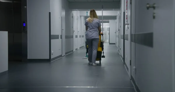 Adult nurse pushes cleaning trolley down medical center hallway. Health worker goes to hospital ward for cleaning. Cleaner takes care of cleanliness of clinic. Medical staff at work in modern clinic.