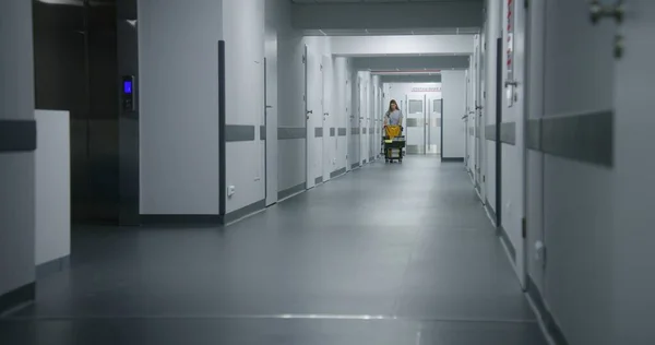 Cleaner goes from hospital room after cleaning. Health worker pushes cleaning trolley down clinic corridor. Nurses take care of cleanliness of medical center. Medical personnel at work in hospital.