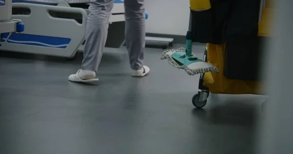 Close up of female health worker mops floor in bright hospital room. Nurse brings cleaning trolley to hospital ward. Cleaner takes care of cleanliness in clinic. Medical staff in medical facility.