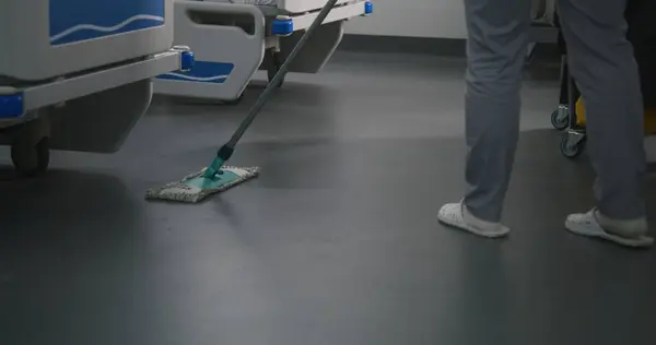 Close up of female health worker mops floor in bright hospital room. Nurse brings cleaning trolley to hospital ward. Cleaner takes care of cleanliness in clinic. Medical staff in medical facility.